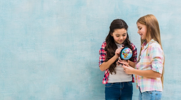 Smiling two girls standing against blue wall looking at small globe