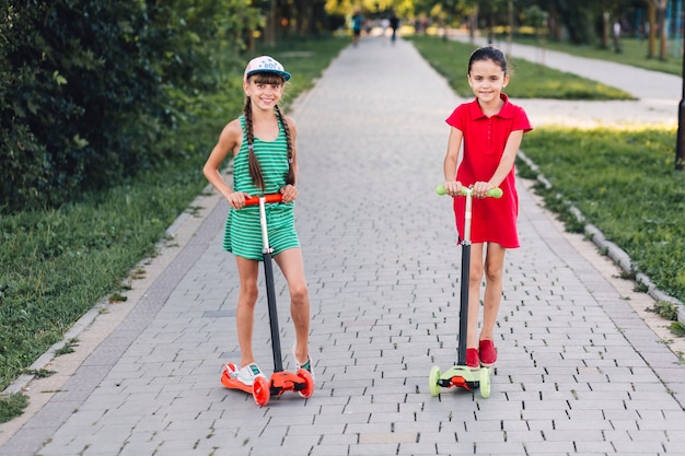 Foto gratuita sorridendo due ragazze che stanno sul motorino di spinta sul passaggio pedonale nel parco