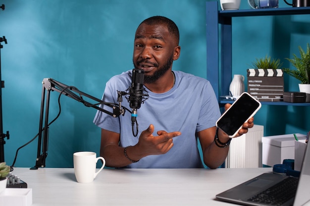 Smiling vlogger holding black screen mockup mobile phone doing online contest in vlogging studio with live setup. influencer hosting online smartphone giveaway while talking into microphone.