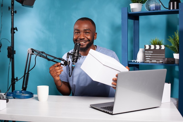 Smiling vlogger holding white box in vlogging studio with professional live setup hosting online giveaway. Influencer showing white mockup famous brand product to fans while talking into microphone.