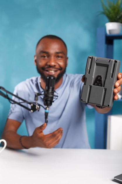 Free photo smiling vlogger presenting camera battery, reviewing videography equipment while recording video. blogger promoting photography gear while live streaming on social media channel