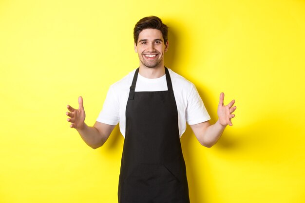 Smiling waiter in black apron holding your logo or box, spread hands as if carry something large, standing over yellow background.