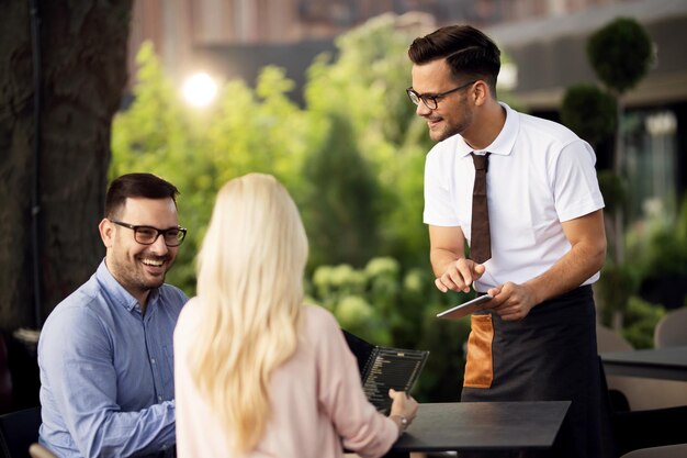 Улыбающийся официант с помощью сенсорной панели во время разговора с гостями и принятия их заказа