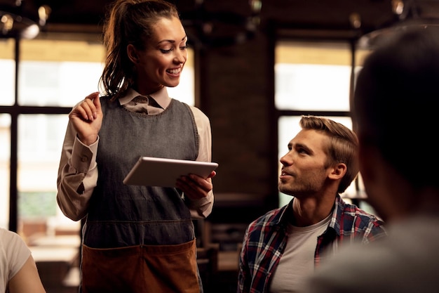 Smiling waitress talking to customers and using touchpad while taking orders in a cafe