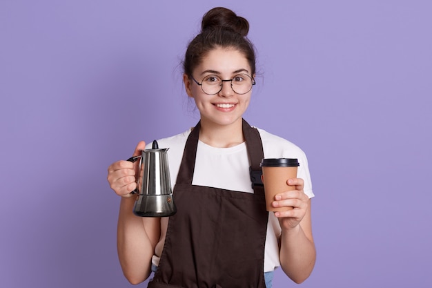 Smiling waitress in white t shirt and brown apron holding pot and take away cup in hands