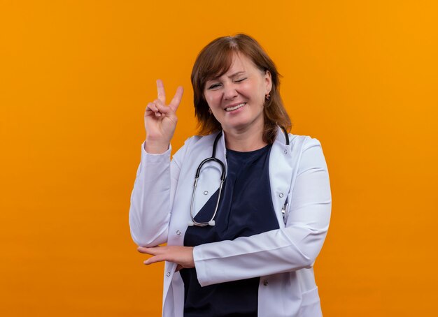 Smiling and winking middle-aged woman doctor wearing medical robe and stethoscope doing peace gesture on isolated orange wall with copy space