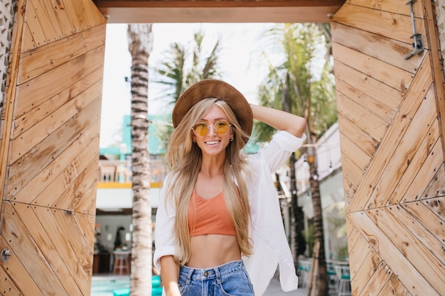 Smiling winsome woman in denim shorts posing with pleasure in summer day. Outdoor portrait of long-haired blonde gorgeous woman in trendy hat laughing in warm morning.