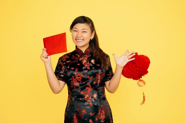 랜턴과 봉투와 함께 웃고. 행복 한 중국 새 해 2020. 노란색 배경에 아시아 젊은 여자의 초상화. 전통 옷을 입은 여성 모델이 행복해 보입니다. Copyspace.