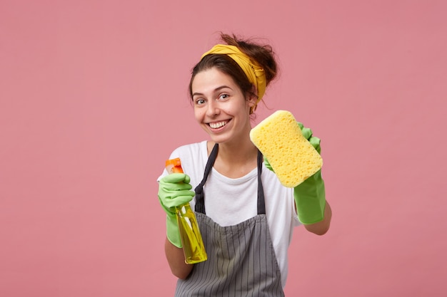 Smiling woman in apron and rubber gloves showing tidy sponge and detergent isolated