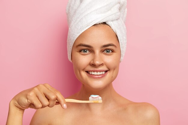 Smiling woman brushes teeth with toothpaste holds wooden toothbrush, wears towel on head, enjoys hygienic procedures