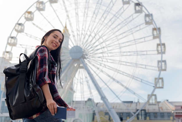 Smiling woman carrying backpack and holding passport and air ticket in front of ferris wheel