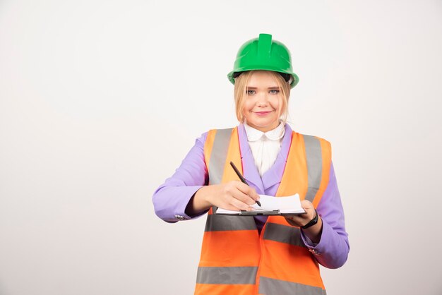 Smiling woman contractor with green helmet holding clipboard on white.