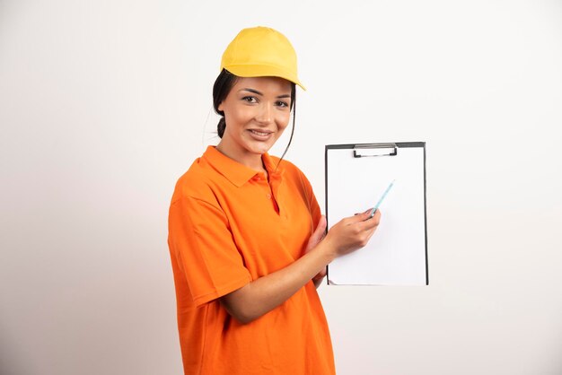 Smiling woman courier showing clipboard on white wall.
