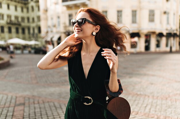 Улыбающаяся женщина в темно-зеленом наряде наслаждается прогулкой по городу