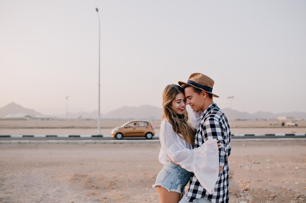 Smiling woman in denim shorts standing with her boyfriens near the road with yellow car. Beautiful young hugging couple of travelers meet sunset at the motorway enjoys nature views