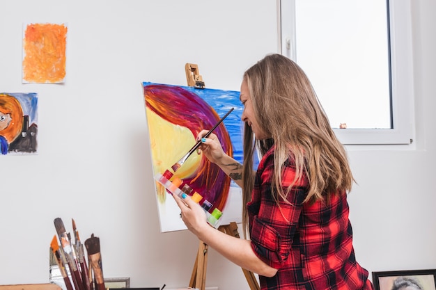 Smiling woman dipping paintbrush into gouache jar standing by easel