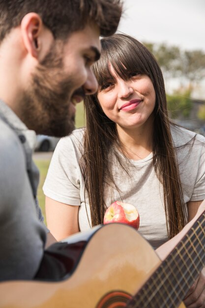 Smiling woman eating apple looking at man playing guitar