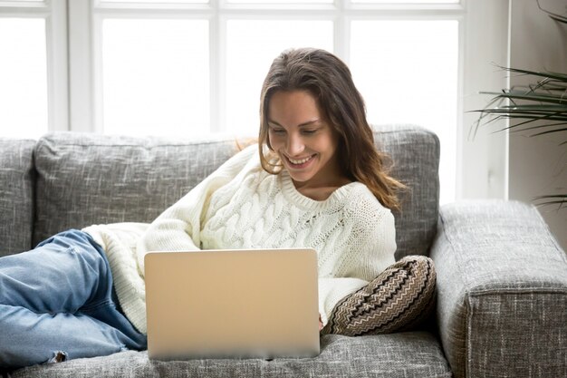 Smiling woman enjoying using laptop relaxing at home on sofa