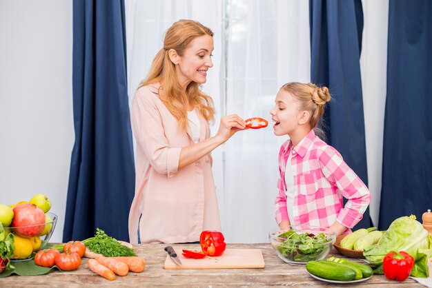 Улыбающаяся женщина кормит ломтик сладкого перца своей дочери