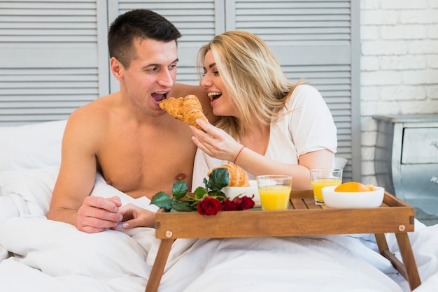 朝食のテーブルの食事の近くのベッドで男性にクロワッサンを与える笑顔の女性