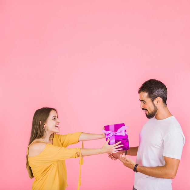 Smiling woman giving pink gift box to her boyfriend standing against pink backdrop