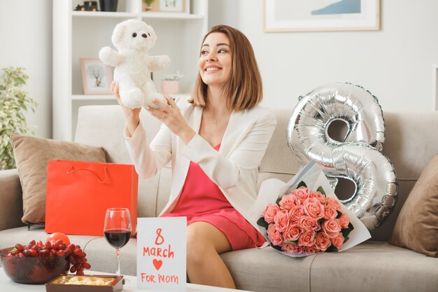 Улыбающаяся женщина в счастливый женский день держит и смотрит на плюшевого мишку, сидящего на диване в гостиной
