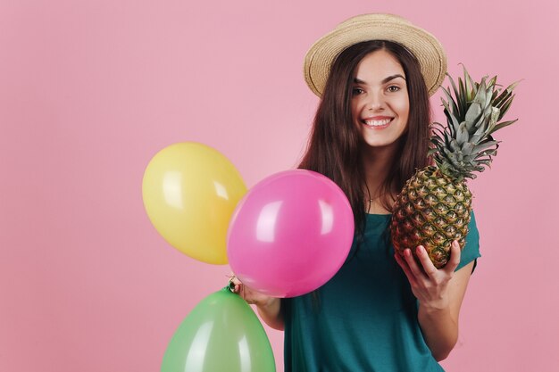 Smiling woman in a hat poses with colorful balloons and a pineapple