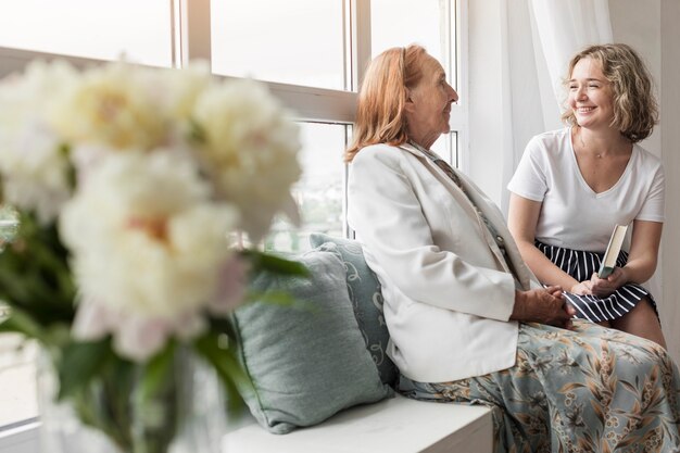 Smiling woman holding book sitting with her granny on window sill