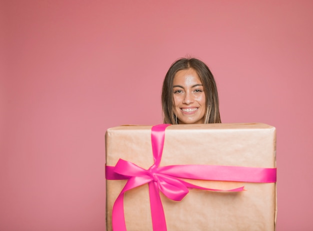 Donna sorridente che tiene il grande contenitore di regalo avvolto con l'arco rosa contro fondo colorato