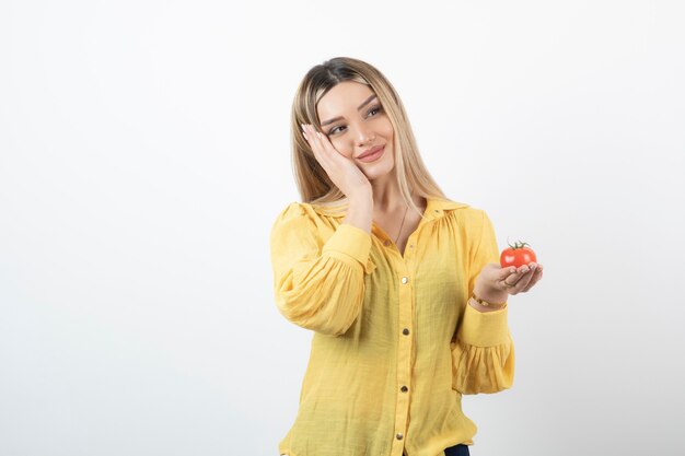 Smiling woman holding red tomato and posing on white wall.