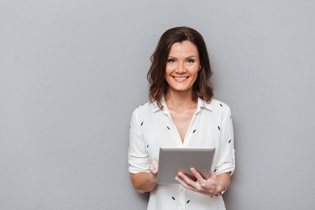 Smiling woman holding tablet computer in hands and looking at the camera on gray