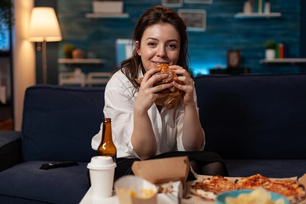 맛있는 버거를 손에 들고 웃고 있는 여성은 테이크아웃 패스트푸드 메뉴가 있는 테이블 앞 소파에 서 있는 텔레비전 코미디 시트콤을 보고 있습니다. 테이크아웃 저녁 식사를 하는 TV 쇼를 보고 있는 행복한 사람.