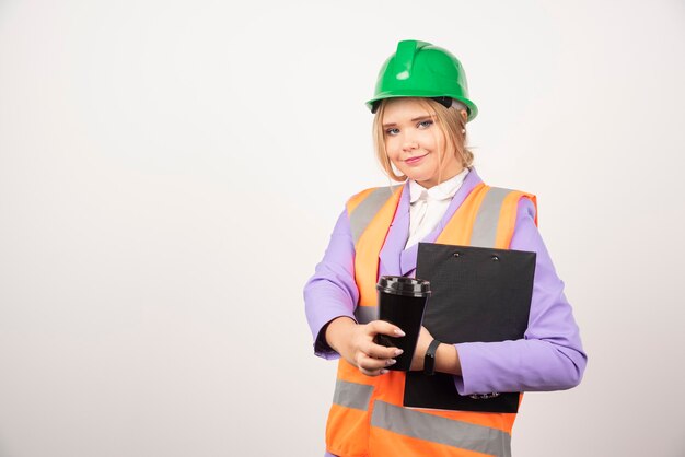 Smiling woman industrial engineer in uniform with clipboard and black cup on white.