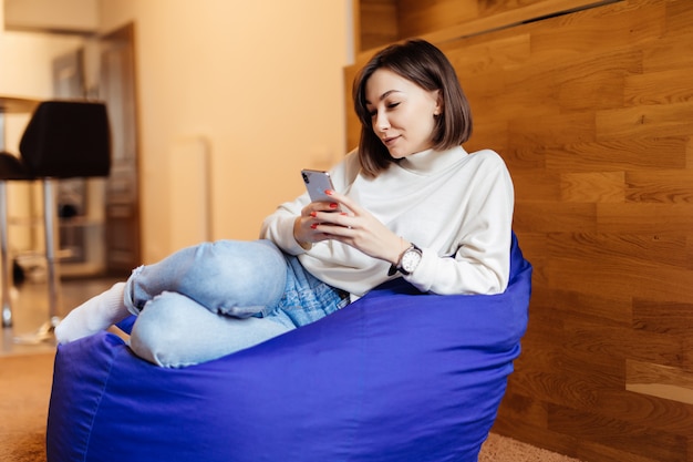 La donna sorridente sta sedendosi nella sedia viola brillante della borsa facendo uso del suo telefono per mandare un sms con i suoi amici