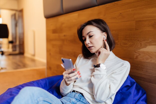 웃는 여자는 그녀의 친구와 문자 메시지에 대한 그녀의 전화를 사용하여 밝은 보라색 가방 의자에 앉아있다