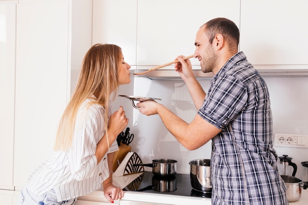 Smiling woman letting man taste a soup with a wooden spoon in the kitchen