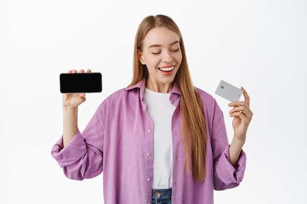 Smiling woman looking happy at her credit card, showing horizontal smartphone screen, recommend application or internet store, standing over white wall