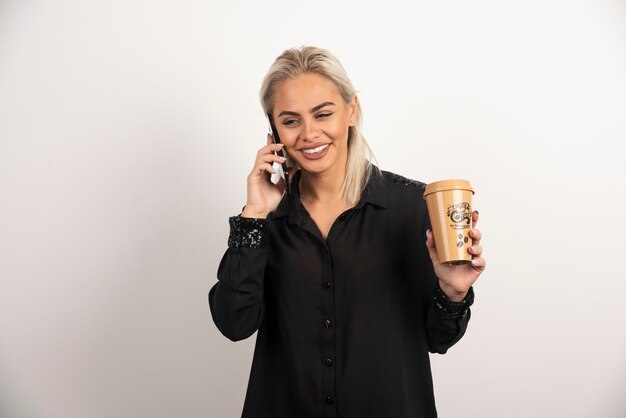 Улыбающаяся женщина смотрит на мобильный телефон и держит чашку кофе. Фото высокого качества