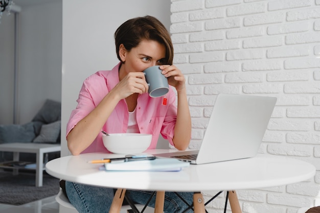 Donna sorridente in camicia rosa che fa colazione a casa a tavola lavorando online sul laptop da casa