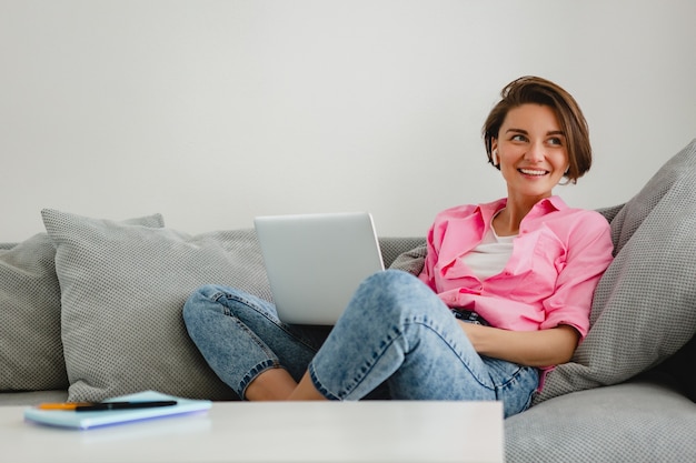 집에서 노트북에서 온라인으로 작업하는 테이블에서 집에서 소파에 편안하게 앉아 분홍색 셔츠에 웃는 여자