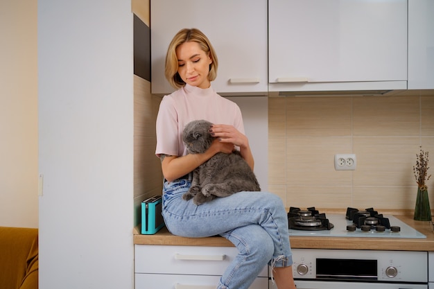 Smiling woman playing with cat in kitchen at home.
