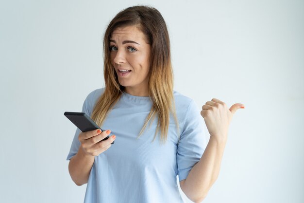 Smiling woman pointing aside with thumb and holding smartphone. 