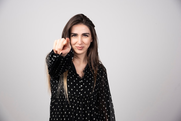 Smiling woman pointing to the camera on a gray wall.