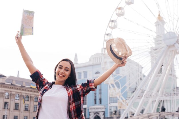 Smiling woman raised her arm with holding map and hat standing near ferris wheel