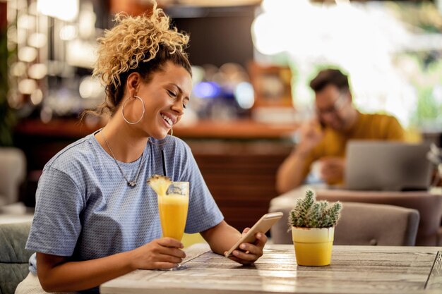 Smiling woman reading text message on cell phone while sitting in a cafe and drinking juice