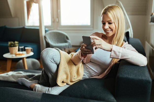 Улыбающаяся женщина отдыхает в своей гостиной и обменивается текстовыми сообщениями на мобильном телефоне.