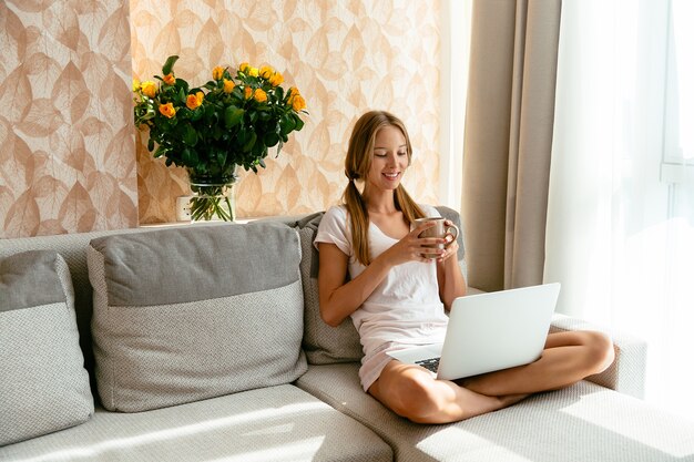 Smiling woman relaxing with cup of tea while watching comedian movie on laptop