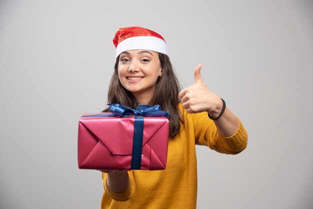 Улыбающаяся женщина в шляпе Санты показывает палец вверх и держит подарочную коробку.
