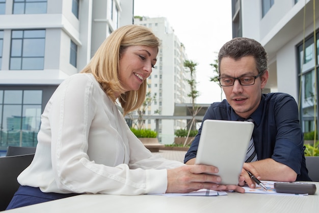 Smiling woman showing male colleague tablet screen in cafe outdoors.