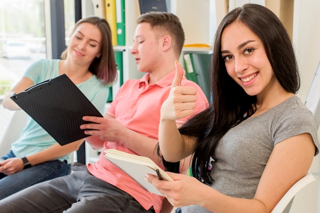 Smiling woman showing thumb up gesture sitting beside her friends holding clipboard